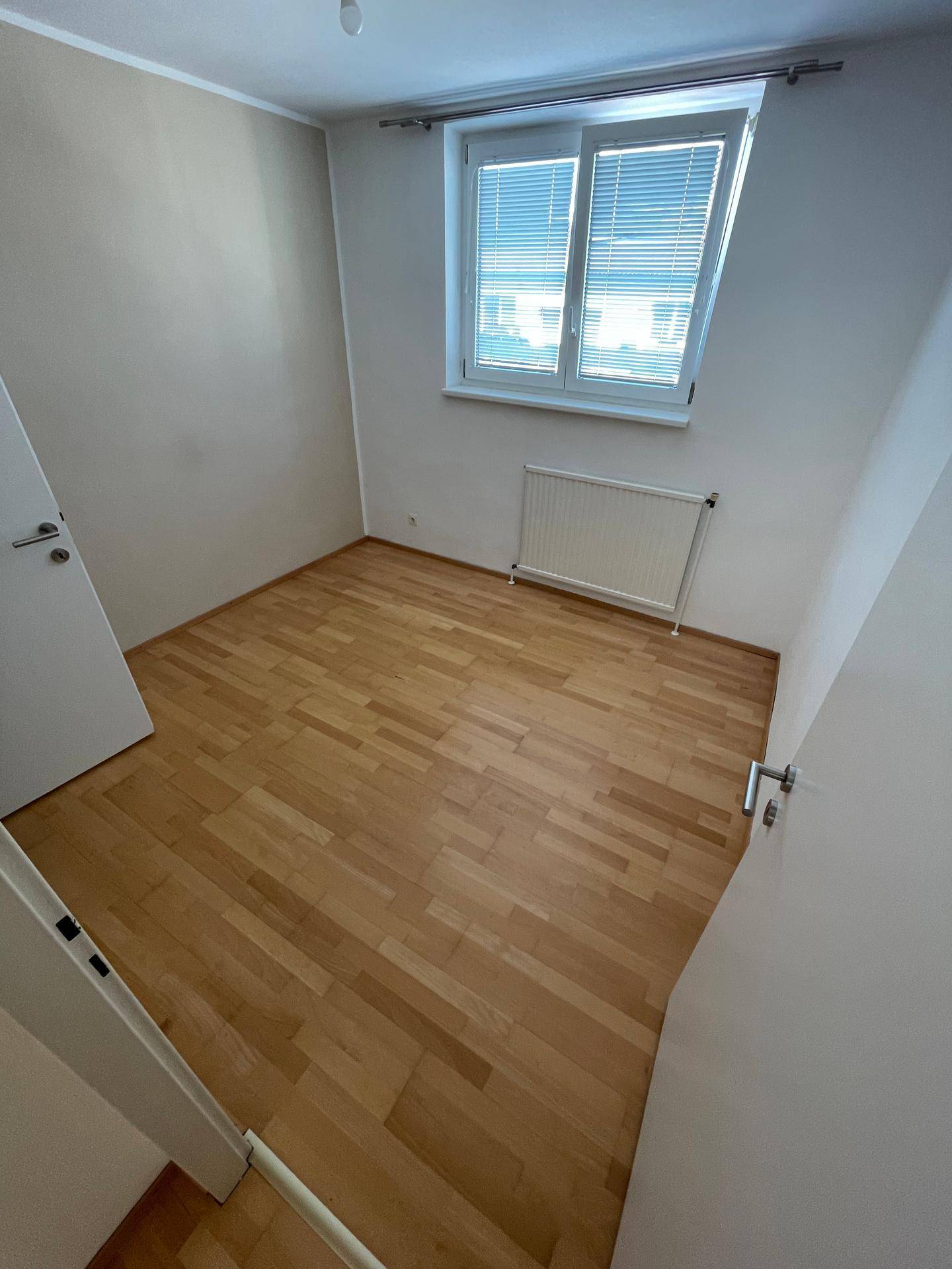 Helles Schlafzimmer mit Parkettboden und Fenster, das viel natürliches Licht hereinlässt.