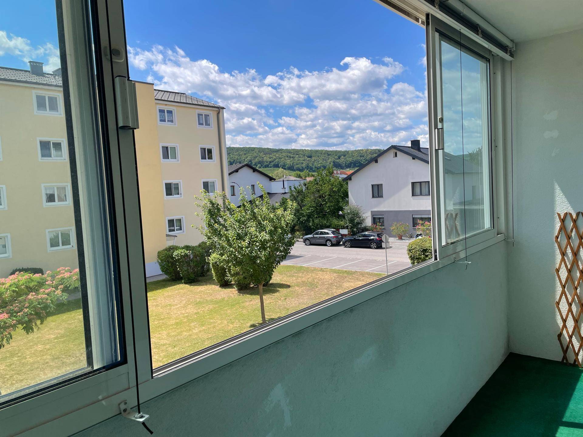 Verglaster Balkon mit Blick auf eine grüne Wohnanlage und Parkplätze unter blauem Himmel.