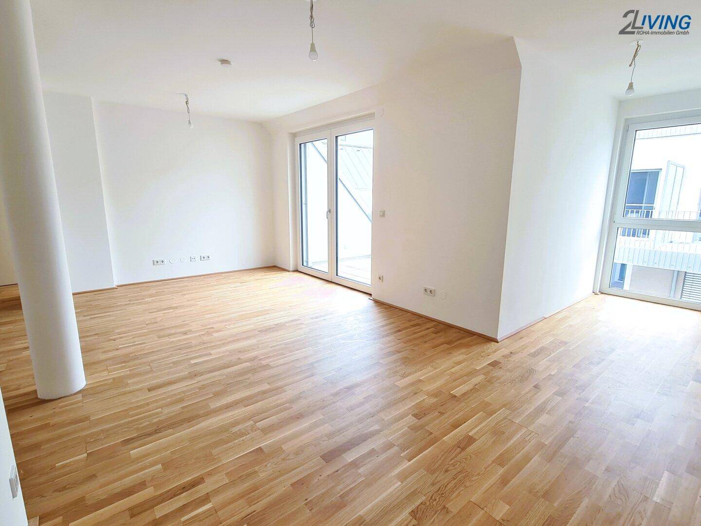 Geräumiger Wohnbereich mit hellem Holzboden und großen Fenstern, die viel Licht hereinlassen.