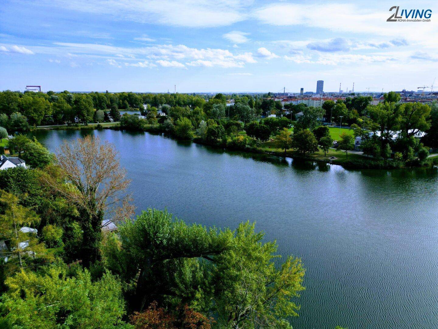 Weitläufiger See umgeben von grünen Bäumen mit einer Stadtkulisse am Horizont unter blauem Himmel.
