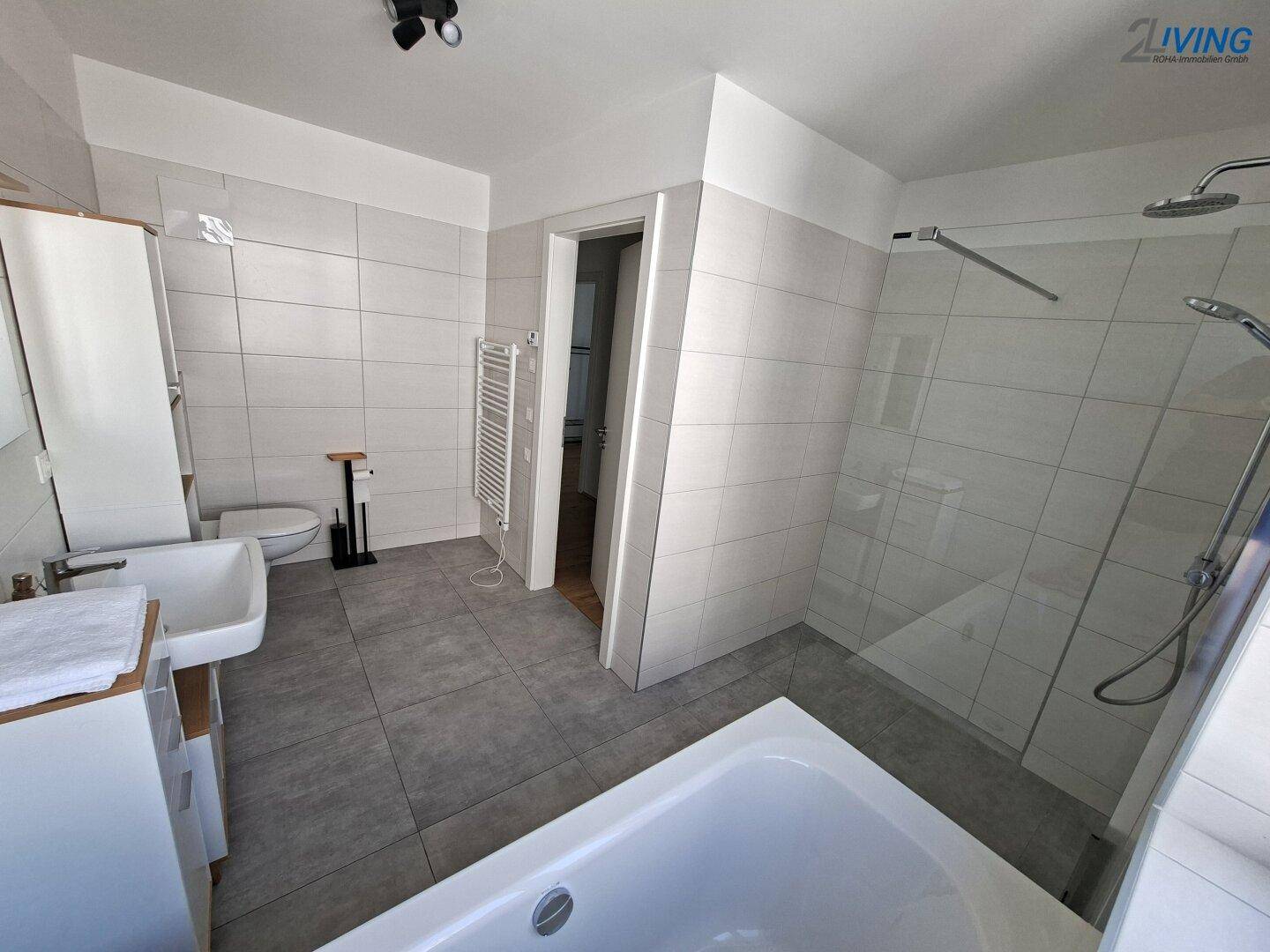 Geräumiges Badezimmer mit ebenerdiger Dusche, WC und beheiztem Handtuchhalter, hell gefliest.