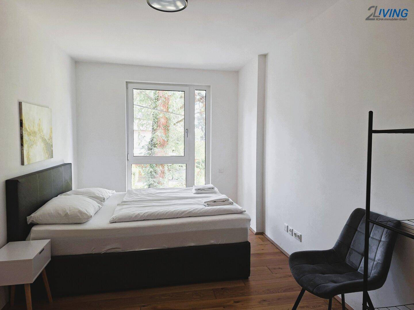Helles Schlafzimmer mit Doppelbett, Holzboden und großem Fenster, das viel Tageslicht hereinlässt.