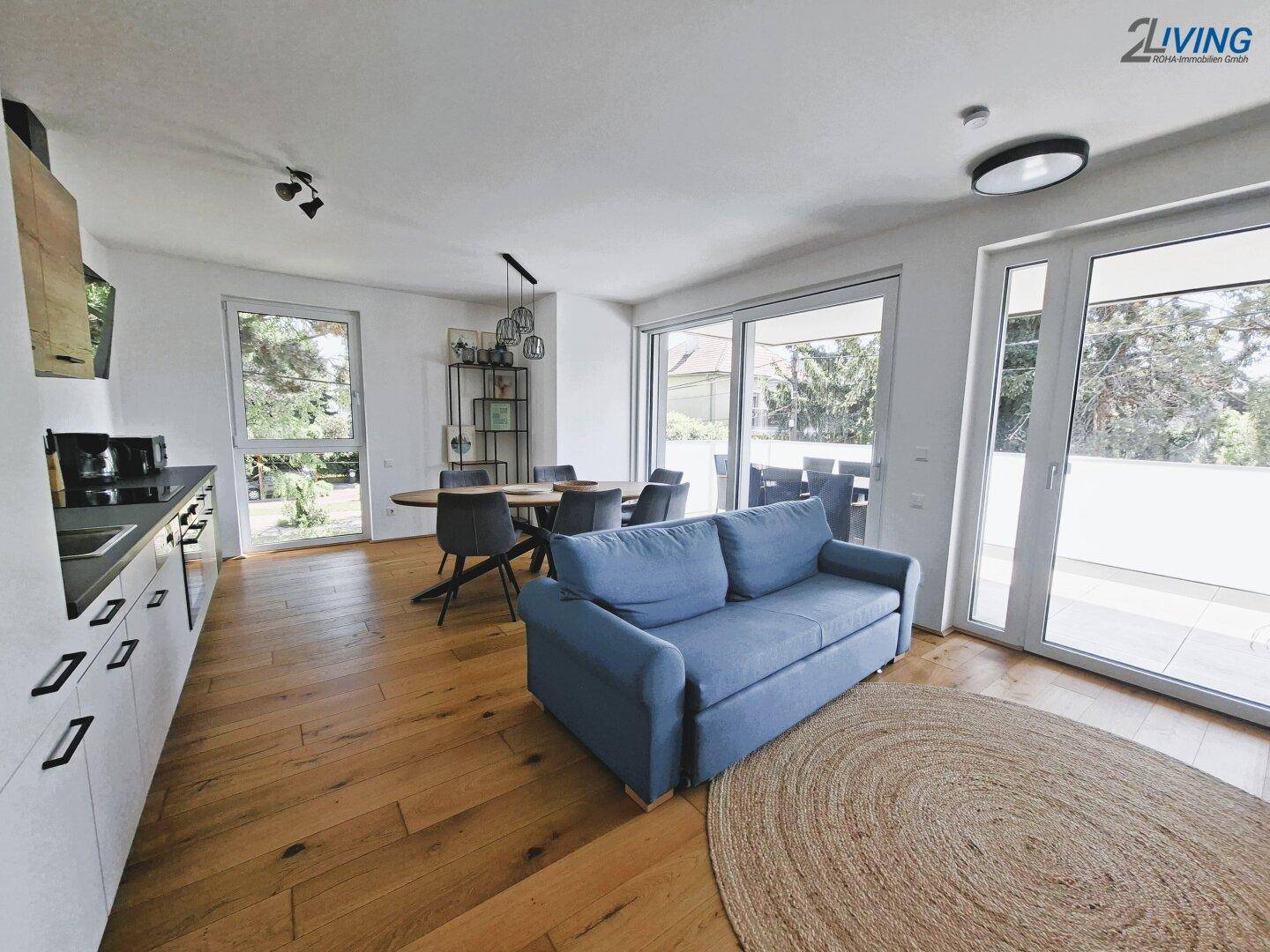 Offener Wohn-Essbereich mit blauer Couch, Holzboden und Zugang zu einem sonnigen Balkon.