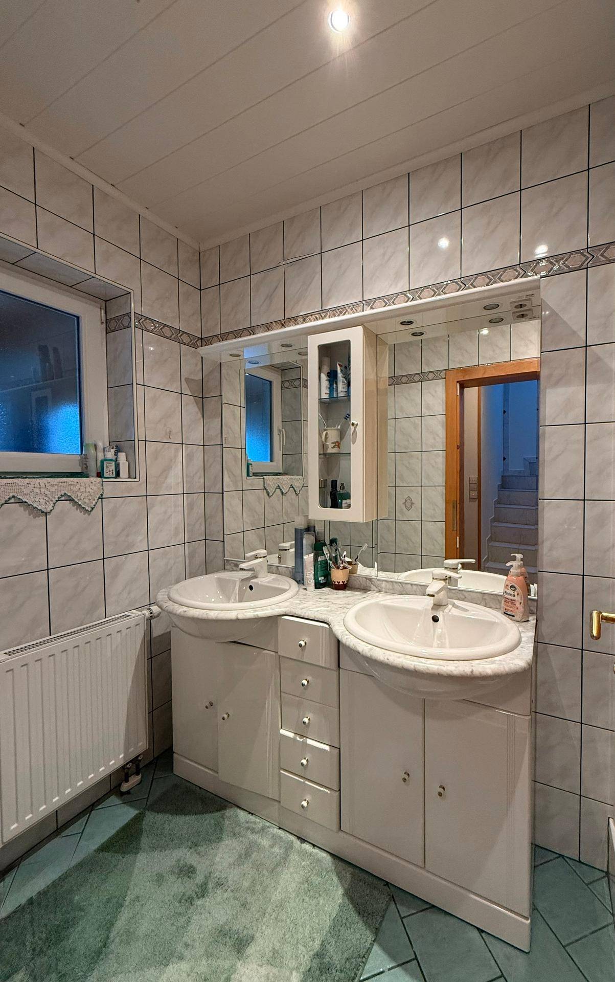 Badezimmer mit Doppelwaschbecken, Spiegelschrank und hellen Fliesen, ausgestattet mit Fenster.