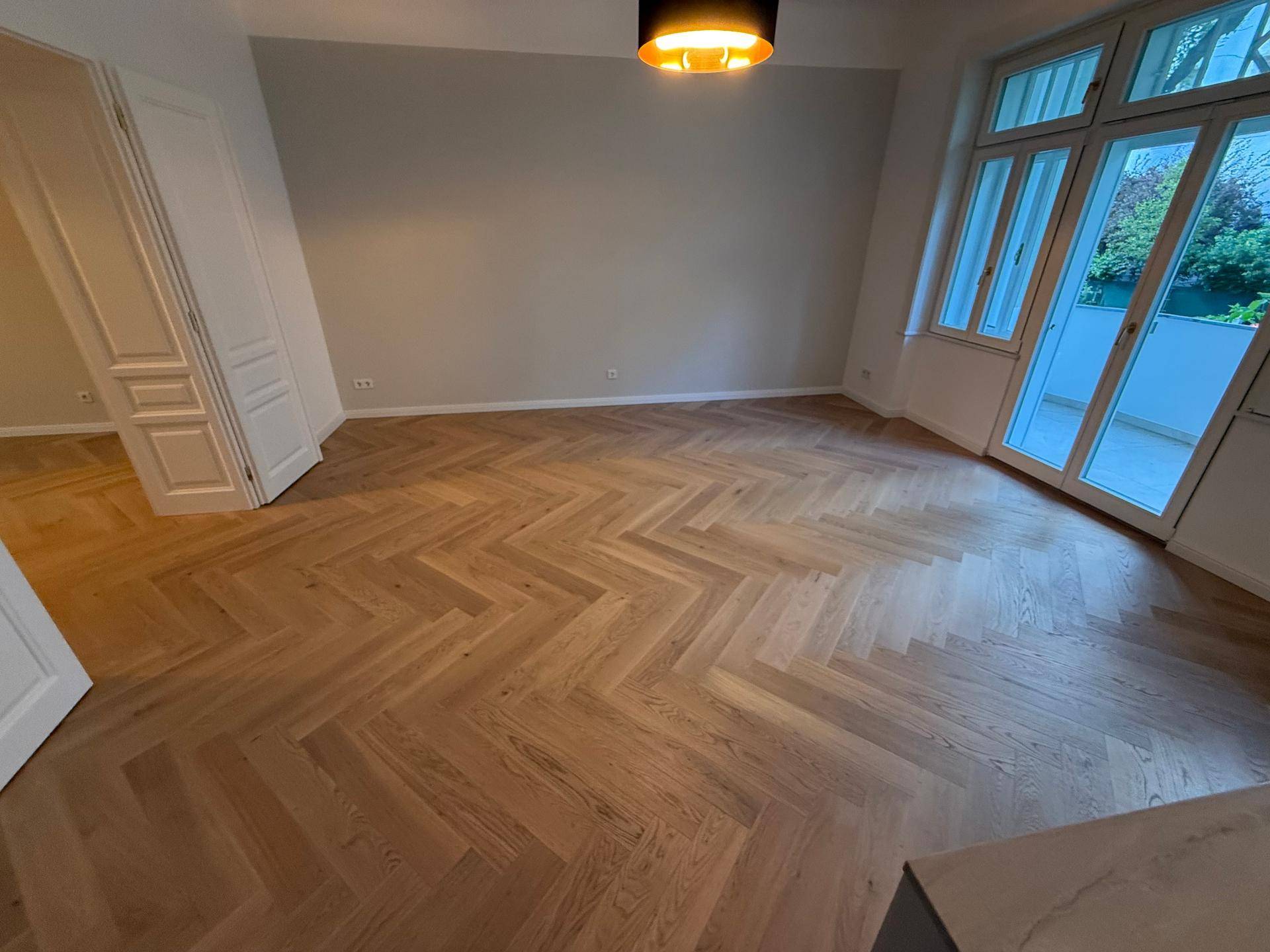 Heller, leerer Raum mit Fischgrätparkett und großen Fenstern mit Balkonzugang.