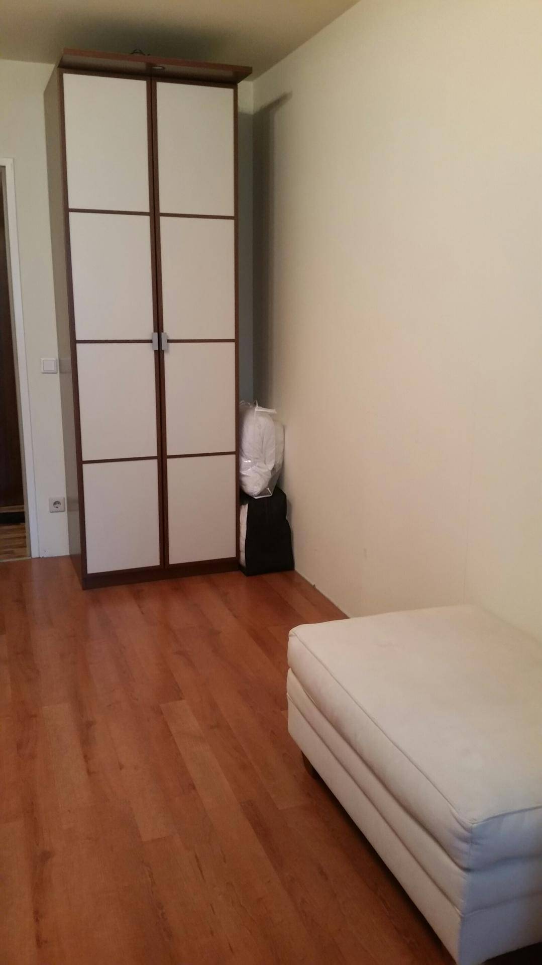 Schlafzimmer mit weißem Kleiderschrank, Polsterhocker und warmem Holzboden.