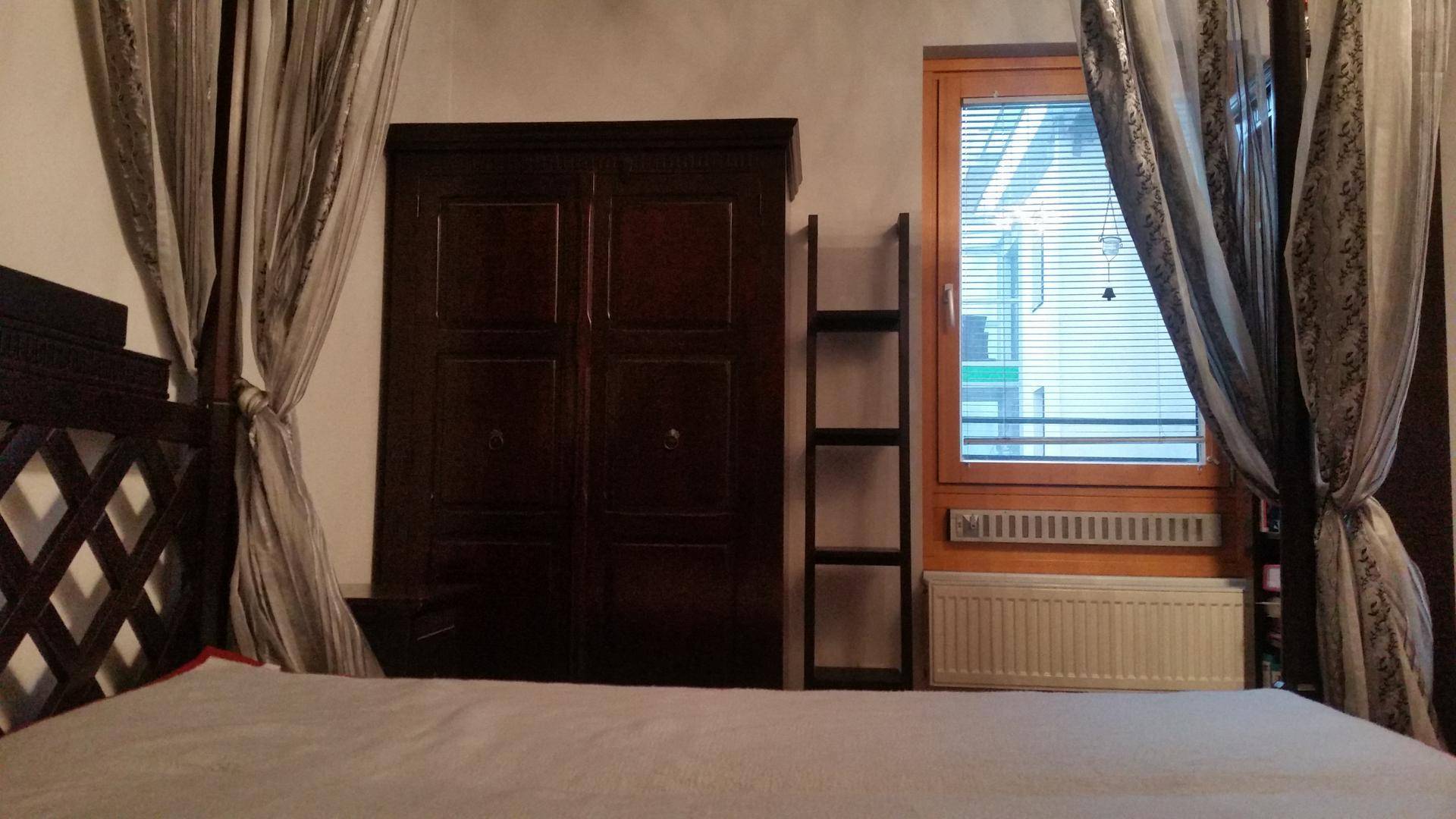 Schlafzimmer mit dunklem Kleiderschrank, Fenster mit Jalousien und Heizkörper.