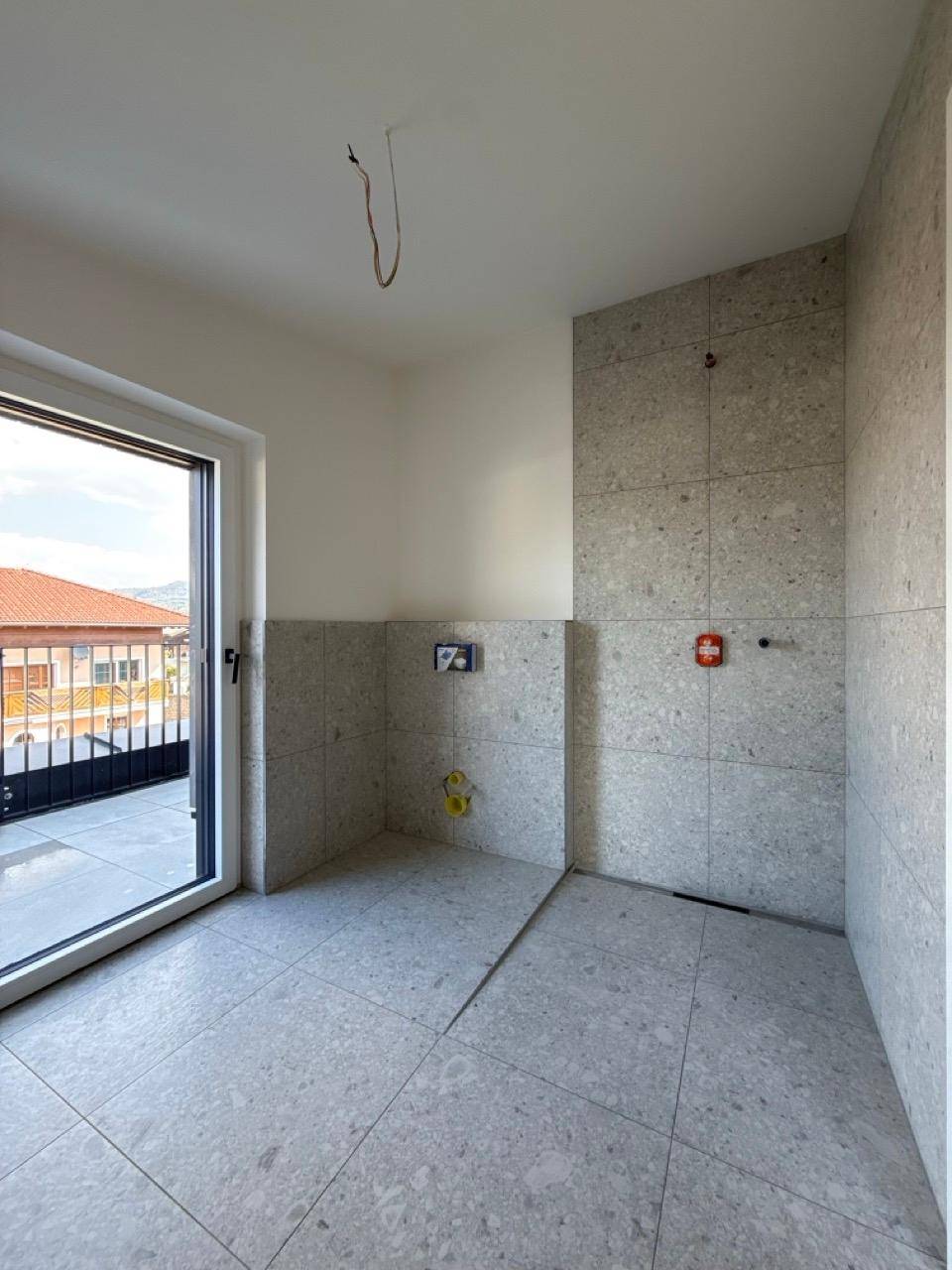 Badezimmer im Bau mit grauen Fliesen, vorbereiteten Anschlüssen und Balkonzugang.