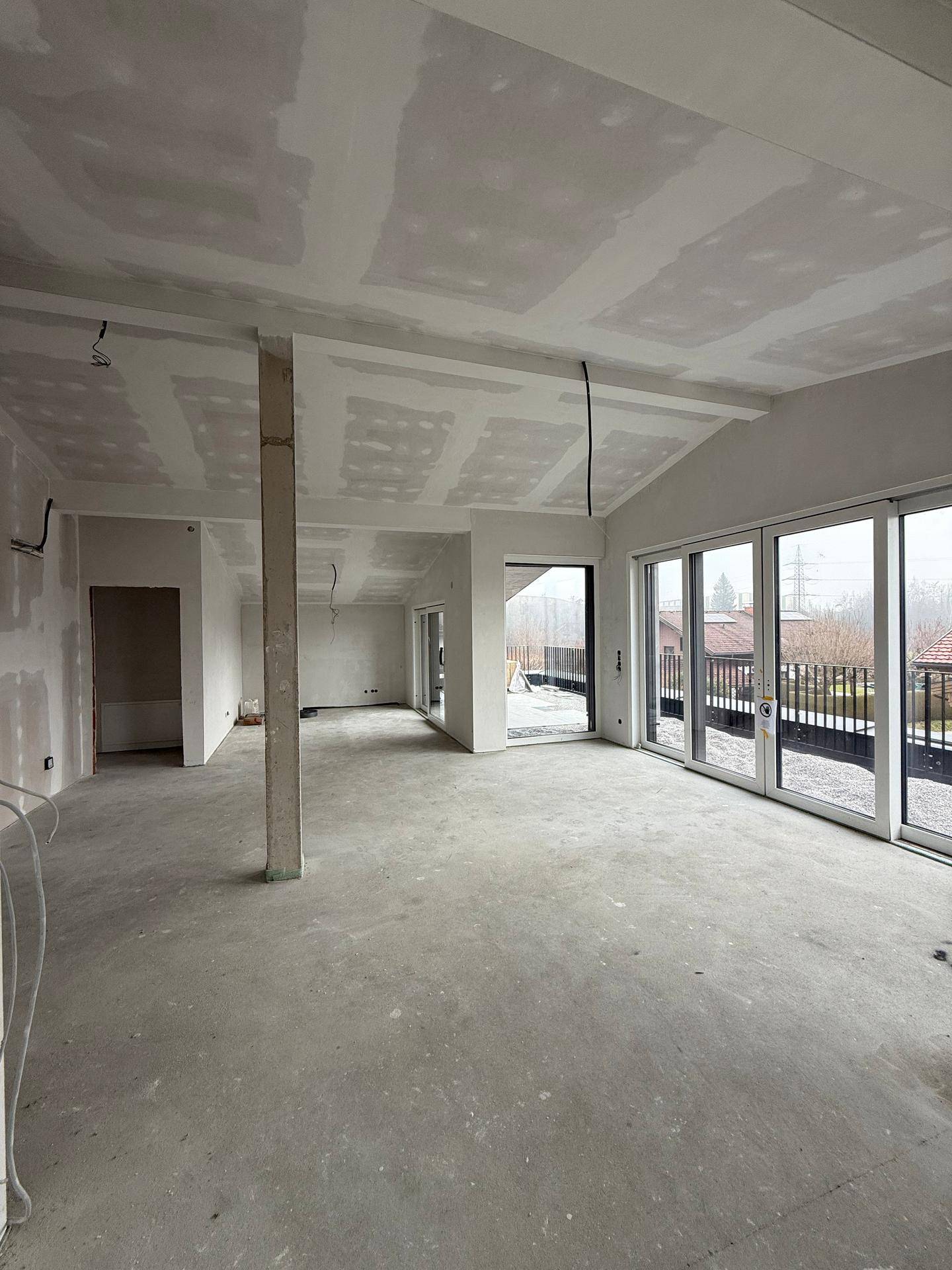 Großer, unfertiger Raum mit Betonboden, schrägen Decken und breiten Schiebetüren zur Terrasse.