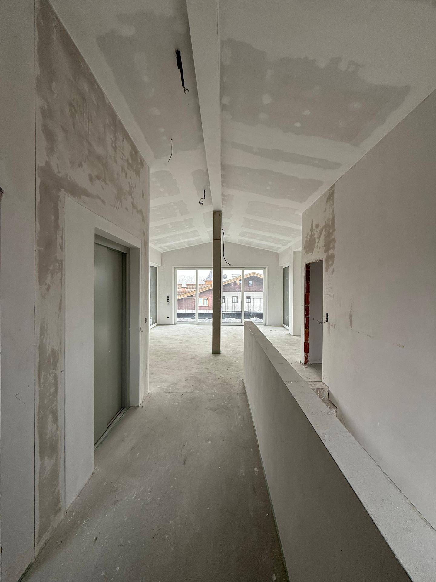 Unfertiger Innenraum mit Trockenbauwänden, schrägen Decken und Blick auf große Fenster.