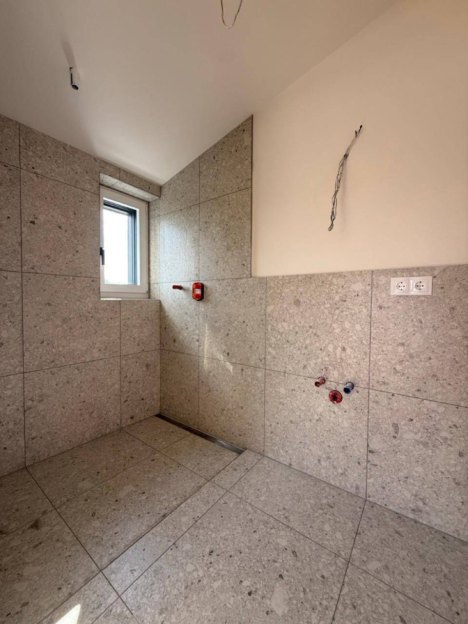 Badezimmer im Bau mit grauen Fliesen, vorbereiteten Anschlüssen und einem Fenster.