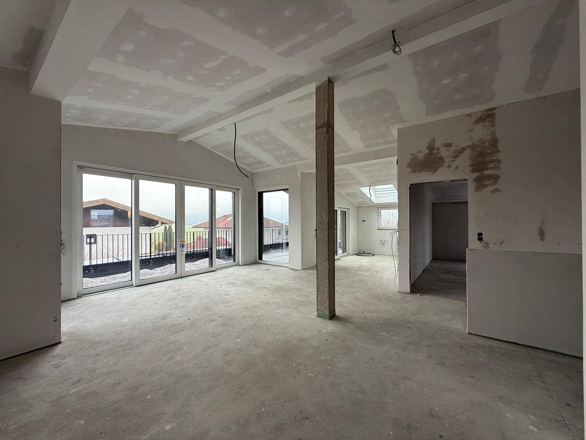 Weitläufiger, unfertiger Raum mit schrägen Decken, Betonboden und großen Fenstern zur Terrasse.