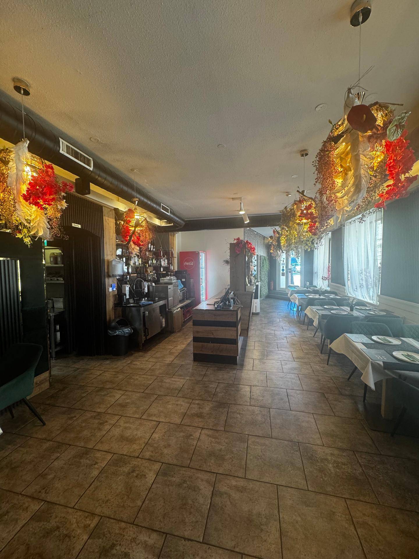 Blick durch den Restaurantraum mit Bar, gedeckten Tischen und Fenstern, die etwas Tageslicht hereinlassen.