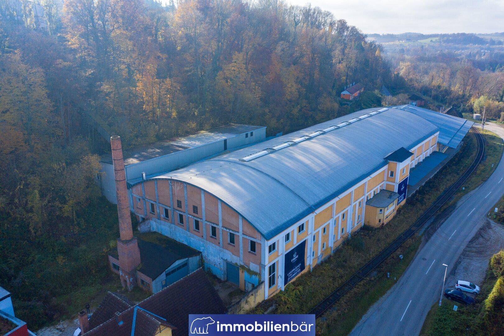 Luftaufnahme eines langen Industriegebäudes mit gewölbtem Dach, hohem Schornstein und umgebenden Bäumen.