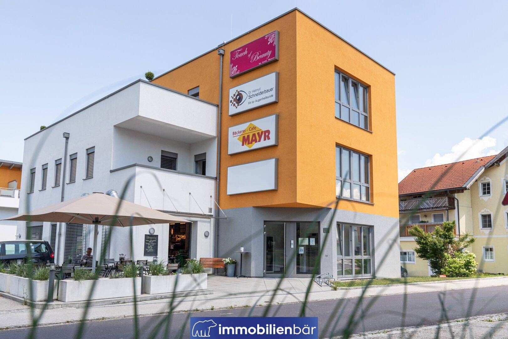 Modernes Geschäftsgebäude mit orangefarbener und weißer Fassade, großen Fenstern und Außensitzbereich eines Cafés.