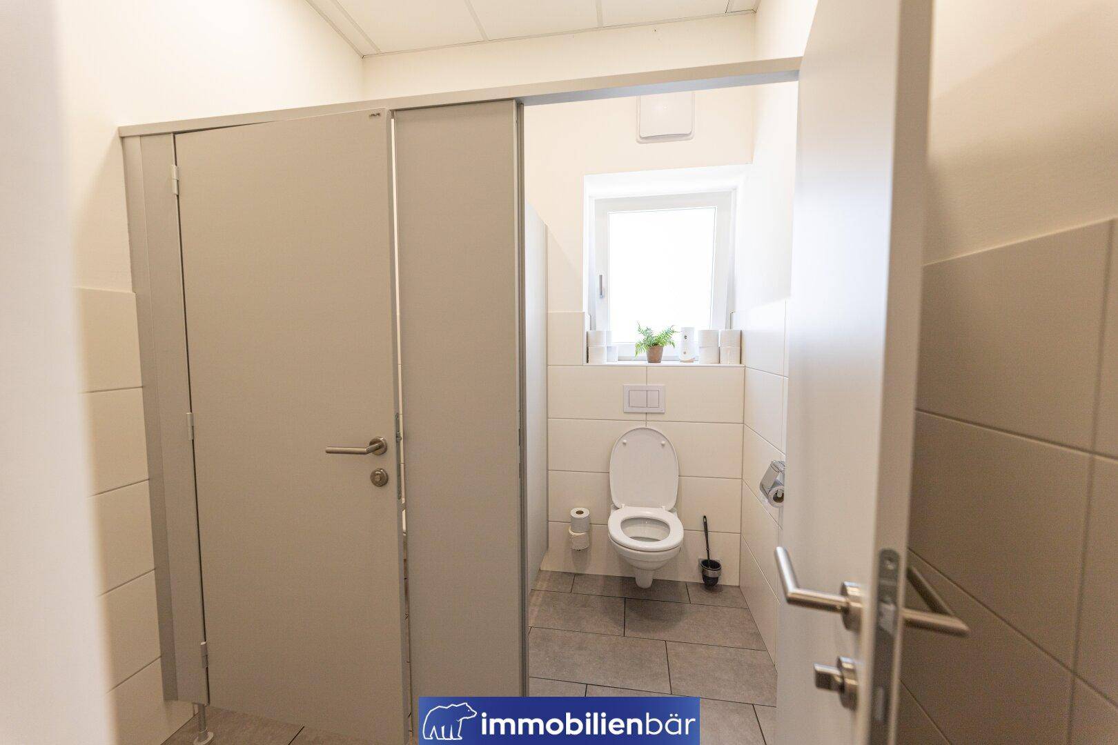 Helle Toilettenkabine mit weiß gefliesten Wänden, Fenster und einfacher Ausstattung in einem Gewerbeobjekt.