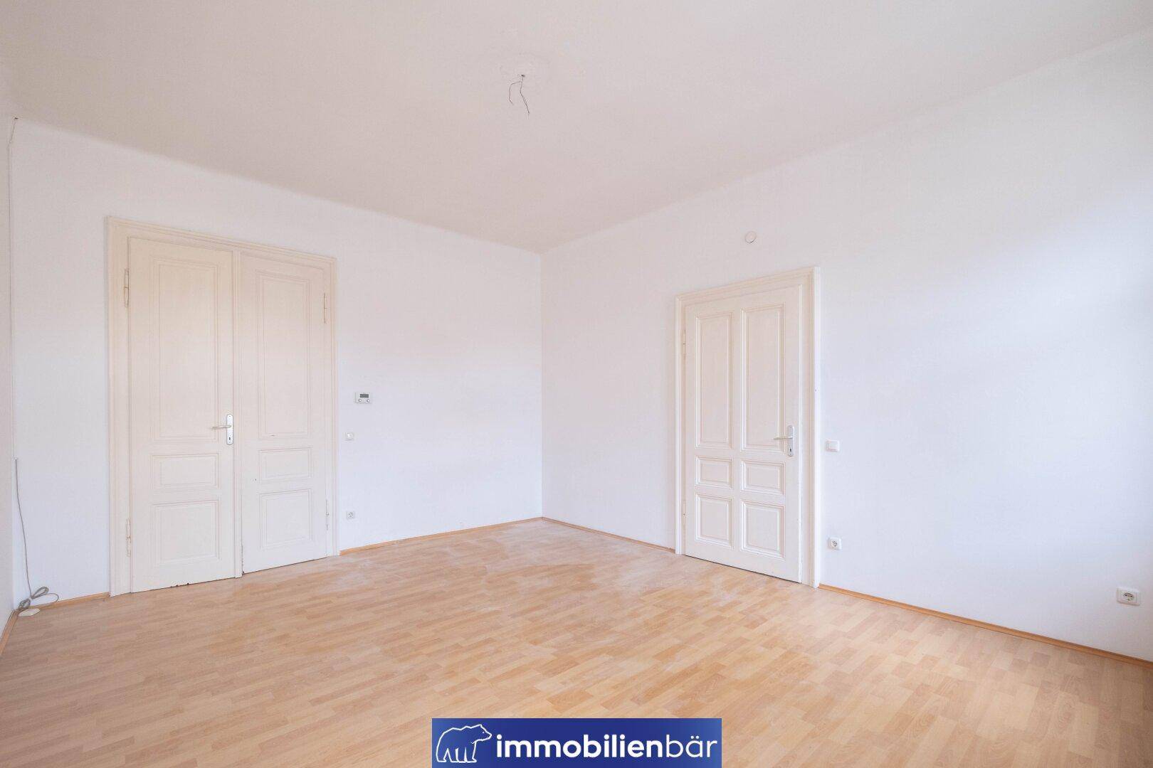 Geräumiges Zimmer mit hellem Parkettboden und zwei klassischen Flügeltüren.