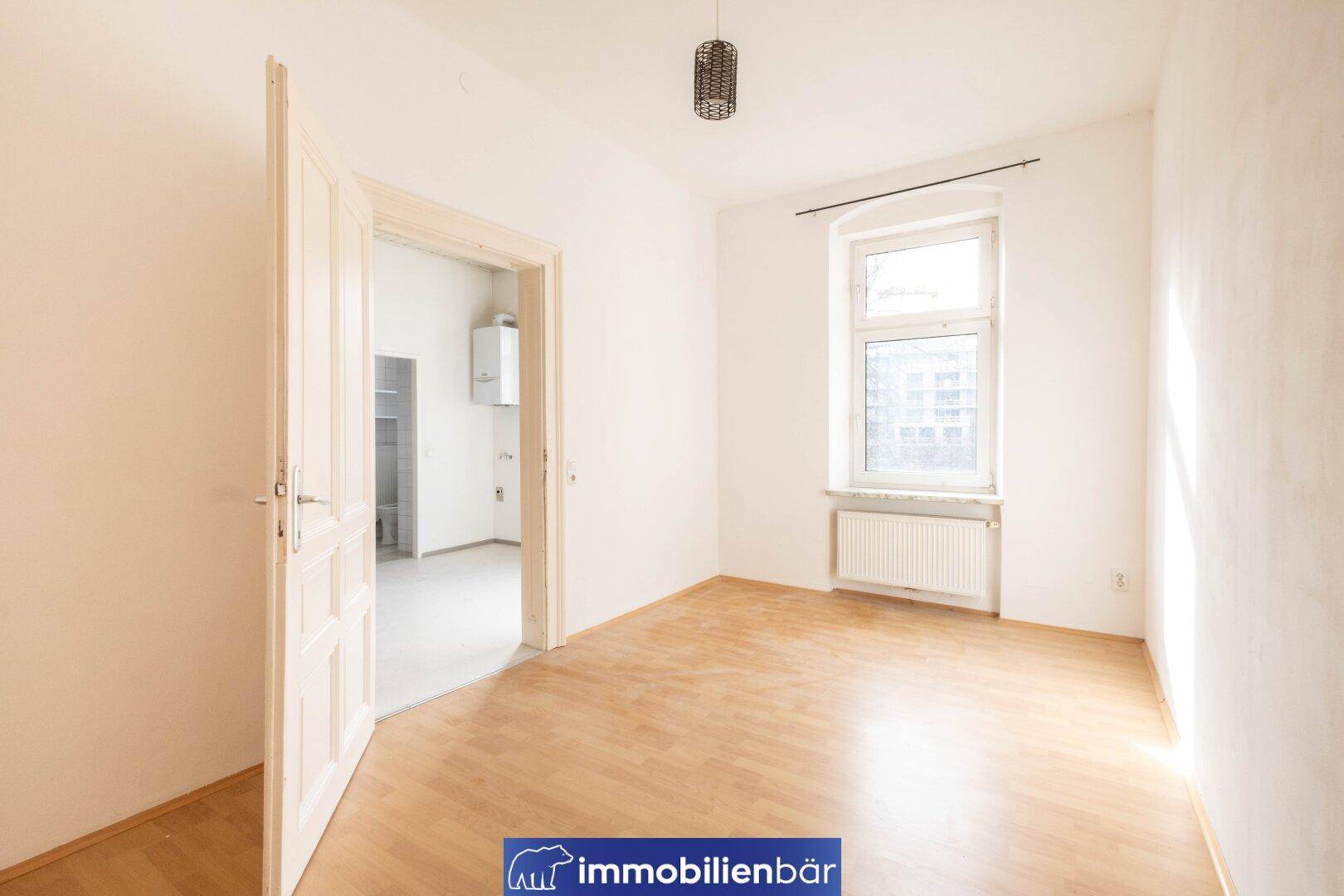 Helles Zimmer mit Parkettboden, großem Fenster und Blick in einen weiteren Raum.
