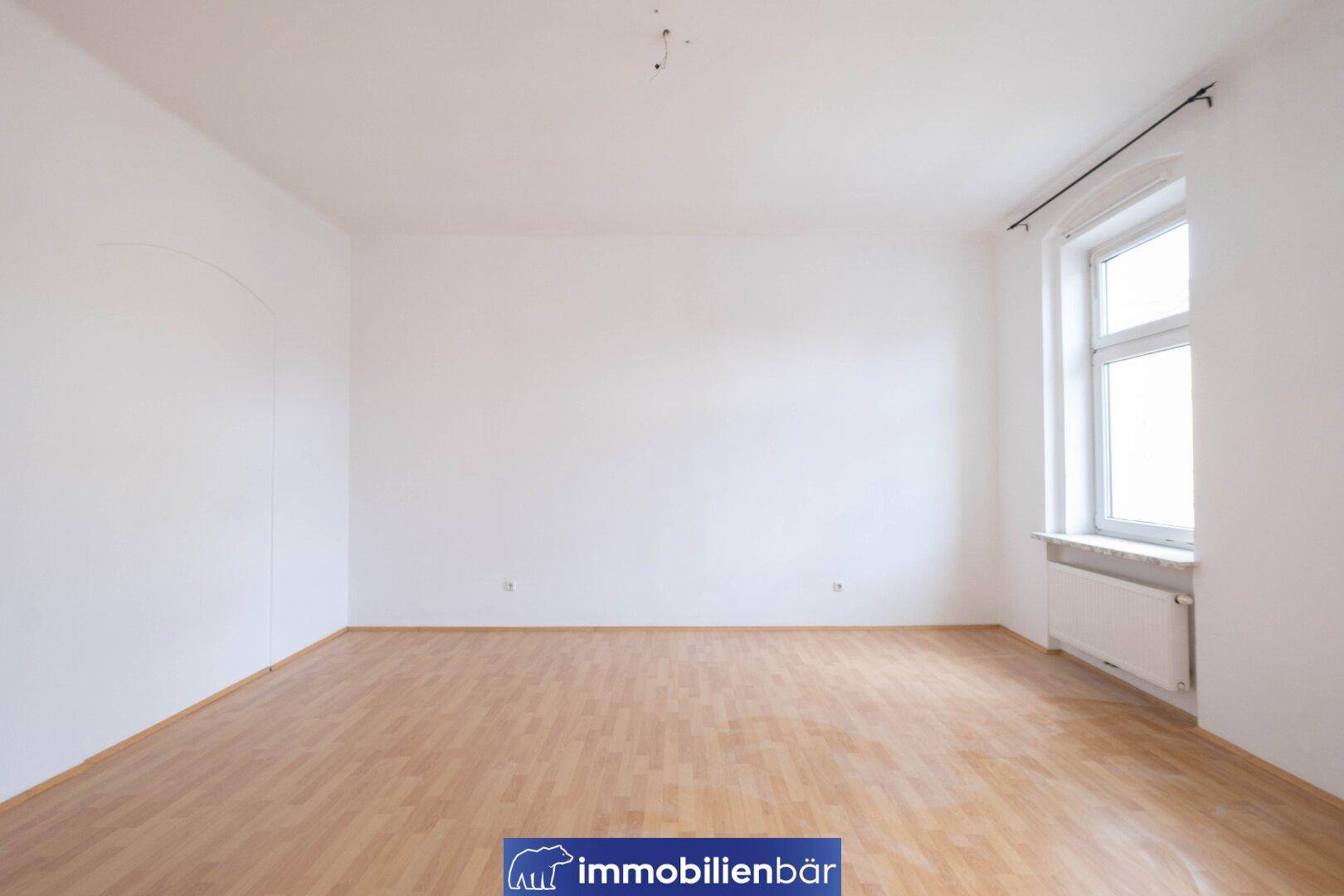 Helles, leeres Zimmer mit Parkettboden und einem großen Fenster für viel Tageslicht.