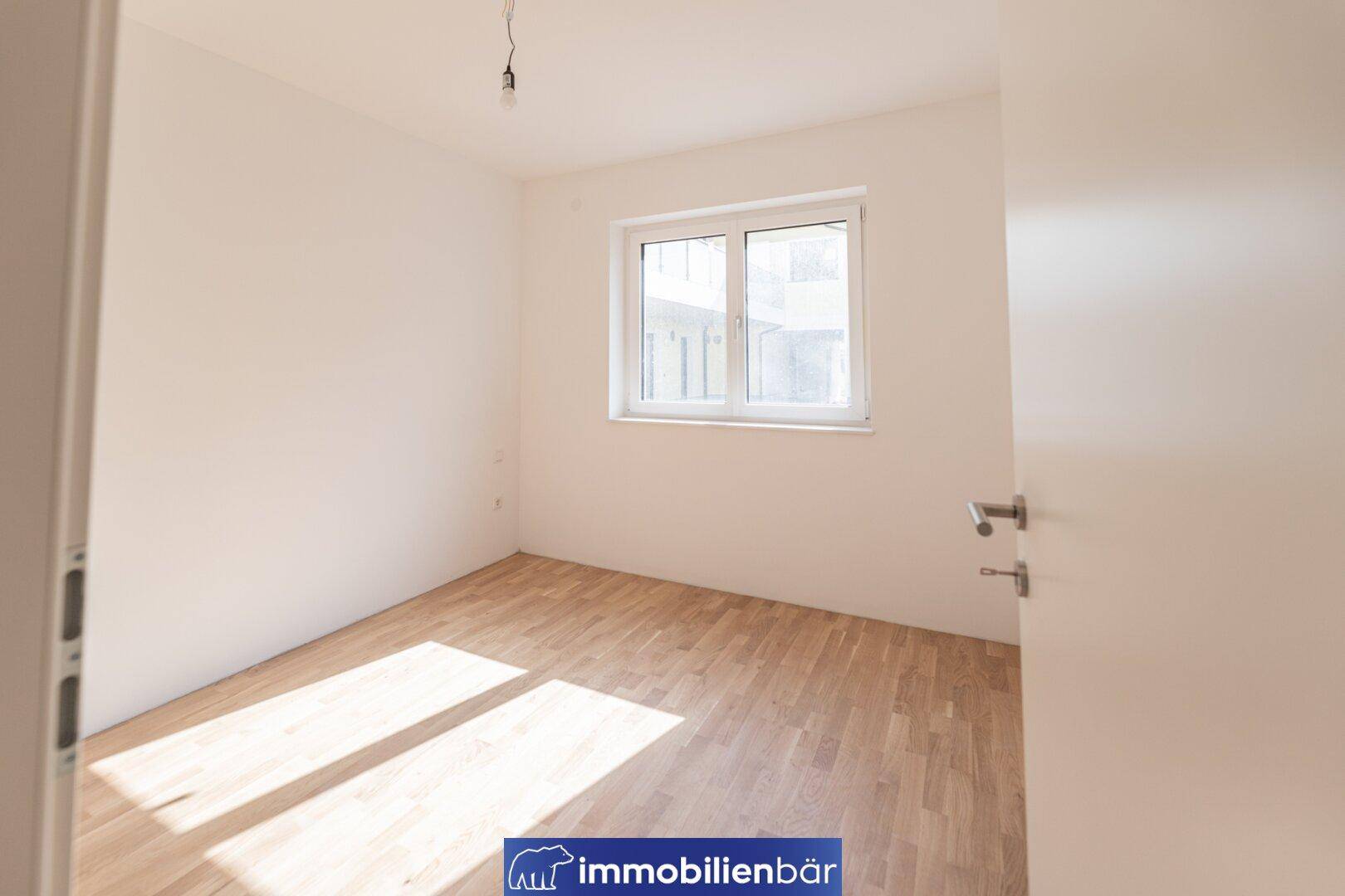Helles, leeres Schlafzimmer mit Parkettboden und einem Fenster, das viel Tageslicht hereinlässt.