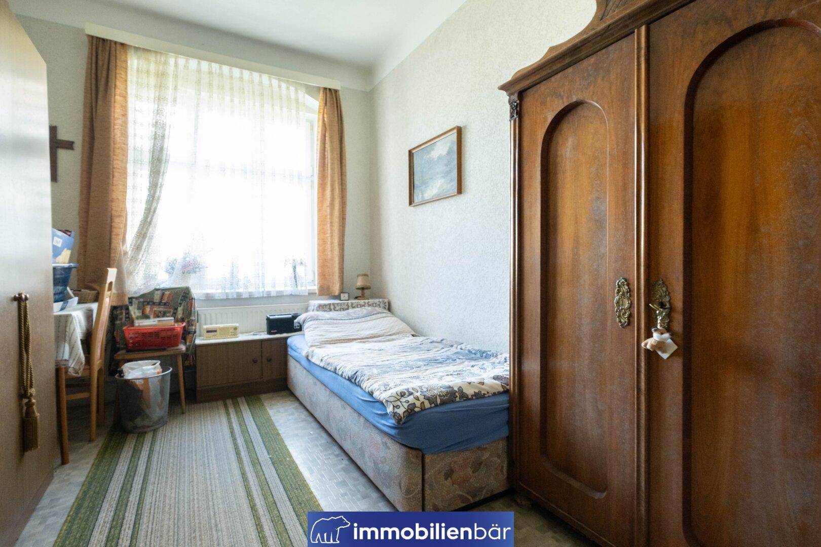 Kleines Schlafzimmer mit Einzelbett, Holzschrank und Fenster mit Vorhängen.