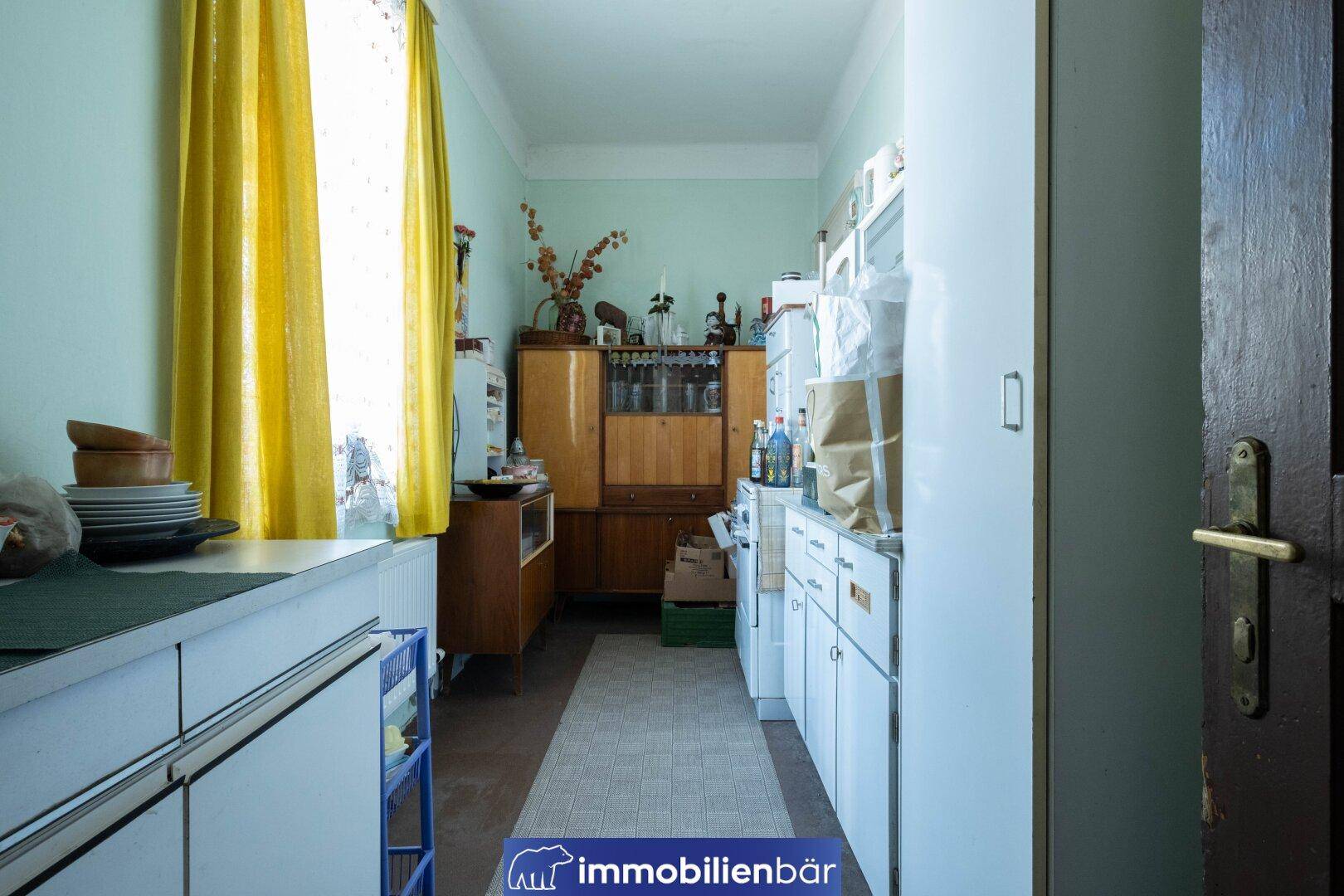 Schmale Küche mit hellgrünen Wänden, weißen Schränken und Fenster mit gelben Vorhängen.