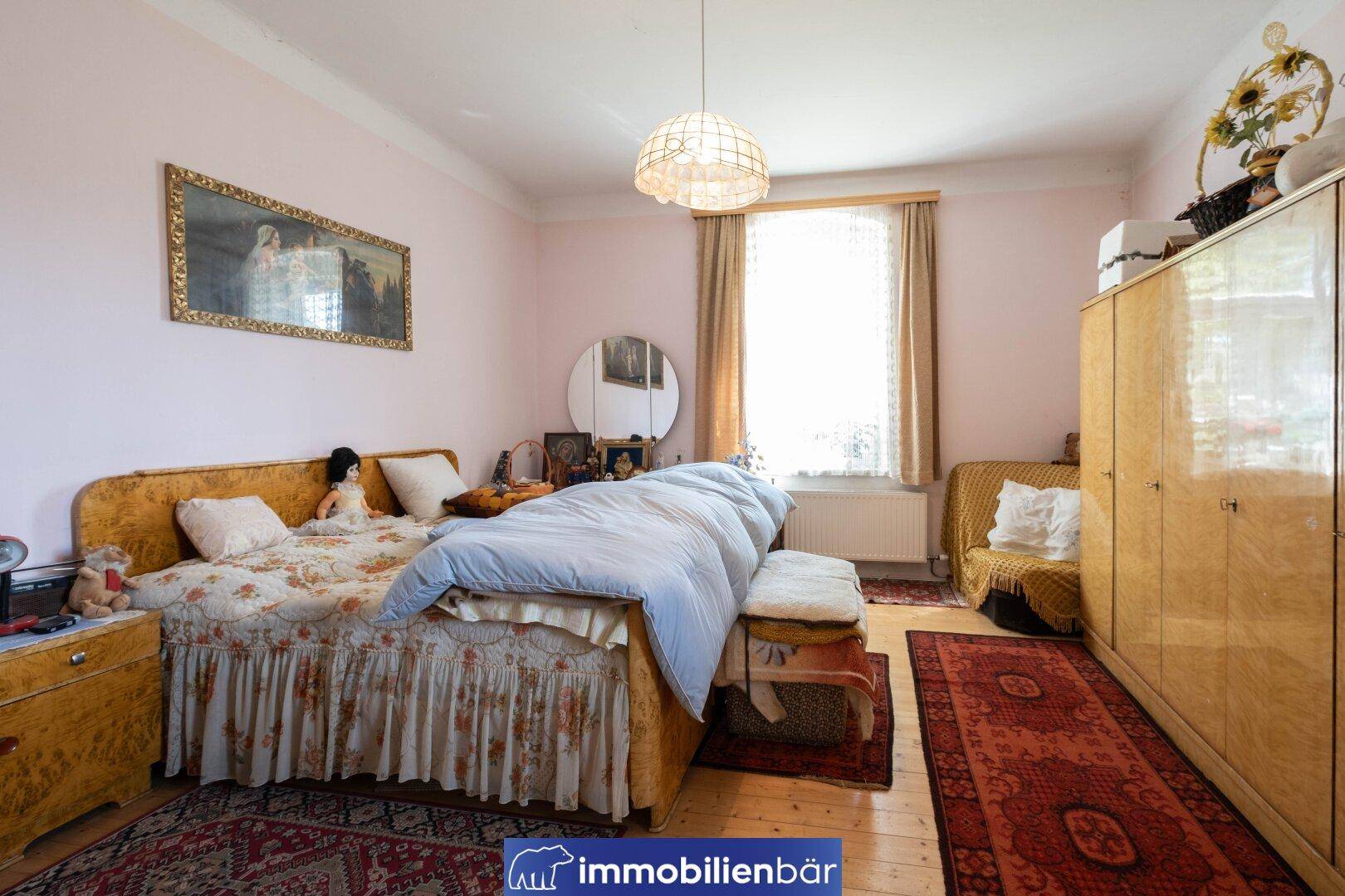 Schlafzimmer mit Doppelbett, Holzmöbeln, Teppich und Fenster mit Vorhängen.