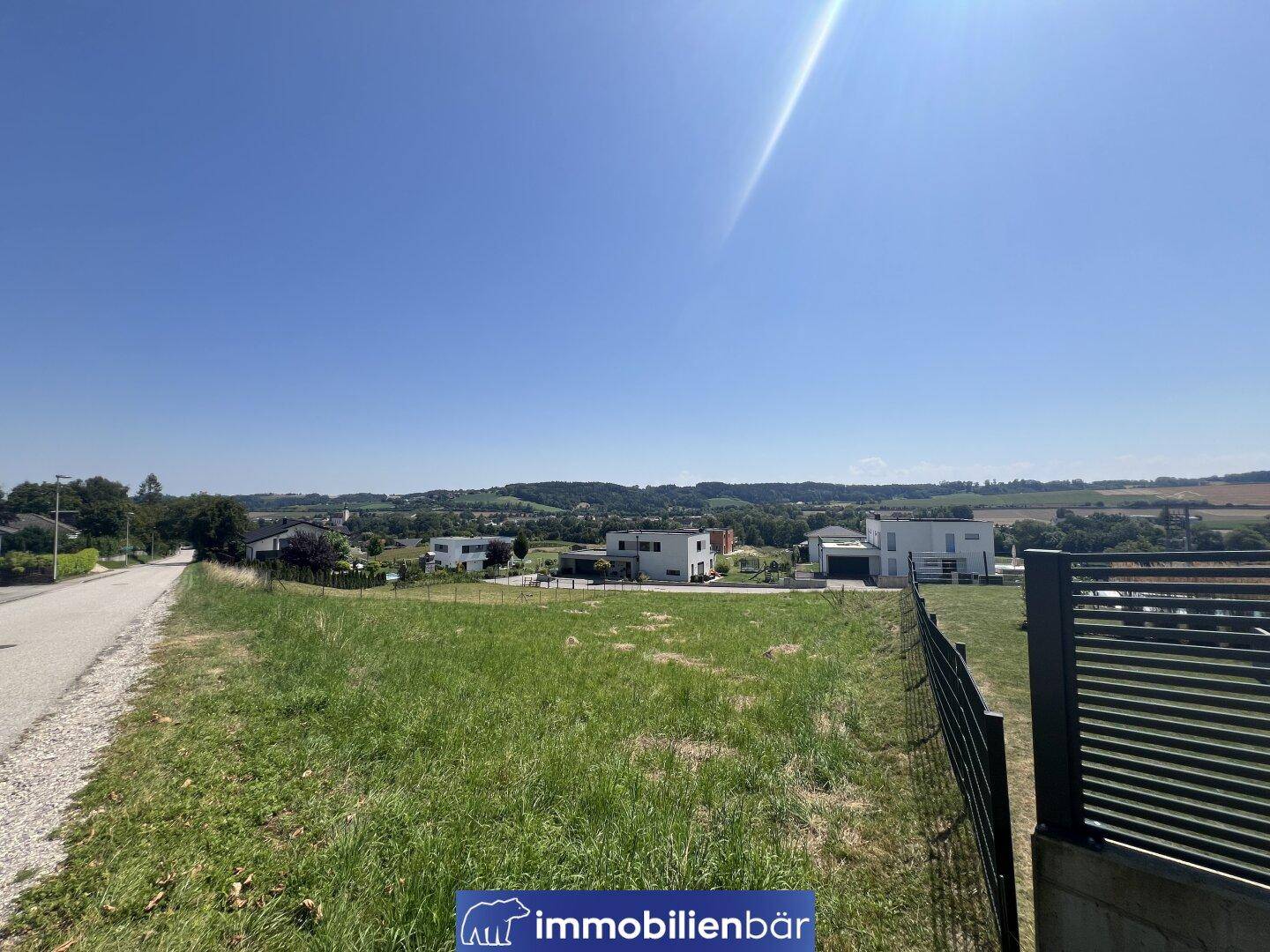 Weitläufiger Blick über ein grünes Grundstück mit Zaun und modernen Gebäuden unter blauem Himmel.