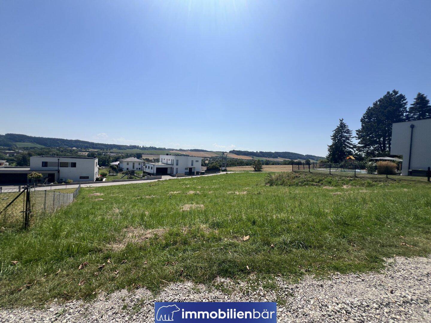 Blick über ein sonniges Wiesengrundstück mit modernen Häusern und hügeliger Landschaft im Hintergrund.