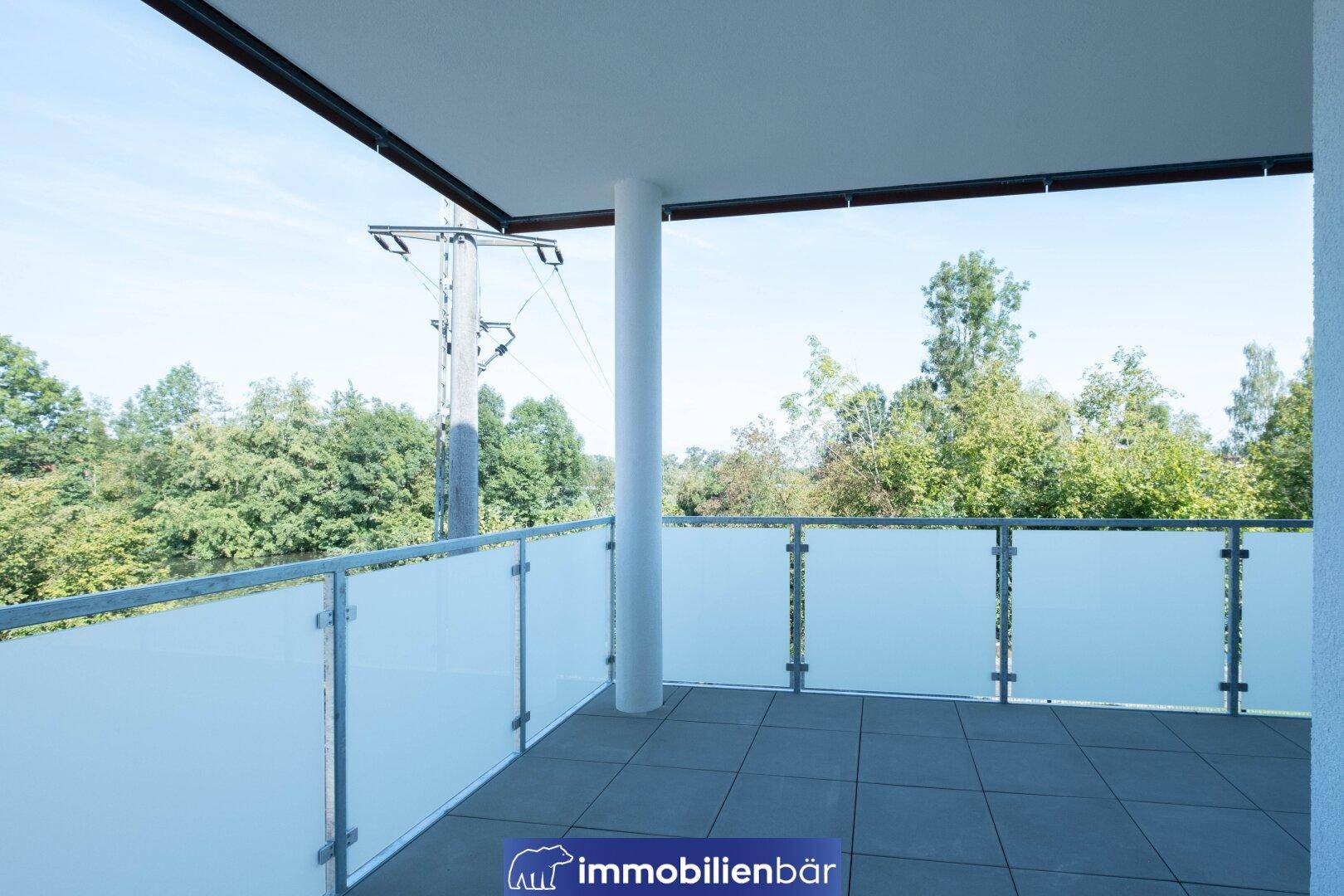 Geräumiger Balkon mit Fliesenboden und moderner Glasbrüstung, umgeben von viel Grün.
