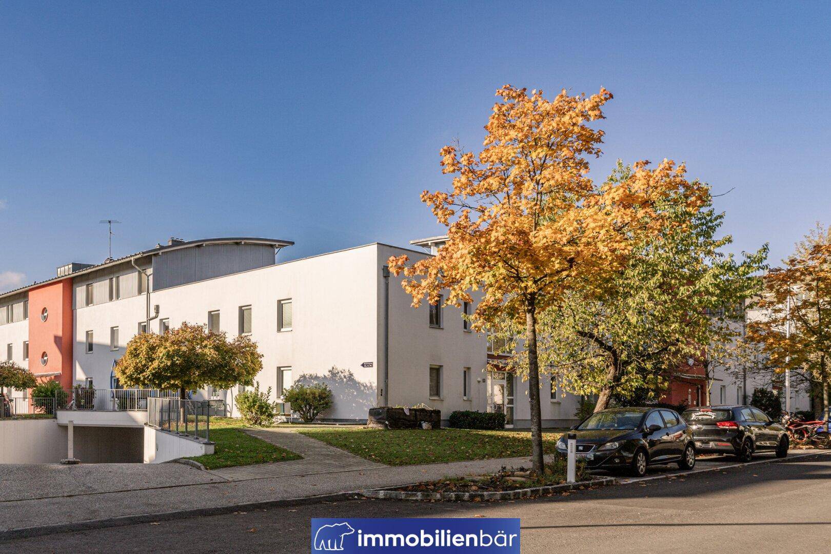 Moderne Wohnanlage mit Tiefgaragenzufahrt, Parkplätzen und herbstlicher Bepflanzung an der Straße.