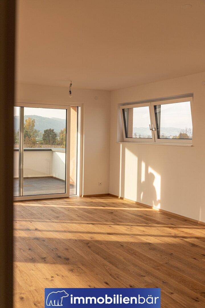 Heller Raum mit Holzboden, Fenster und Fenstertür, die auf eine Terrasse führt.