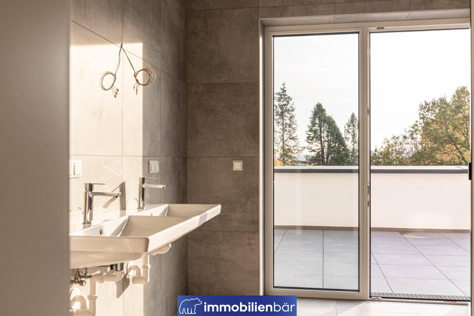 Modernes Badezimmer mit Doppelwaschbecken, grauen Fliesen und Zugang zu einer Außenterrasse.
