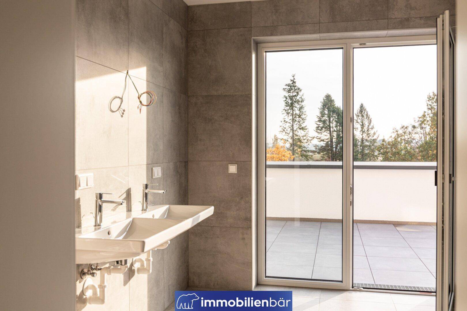 Modernes Badezimmer mit Doppelwaschbecken, grauen Fliesen und Zugang zu einer Außenterrasse.