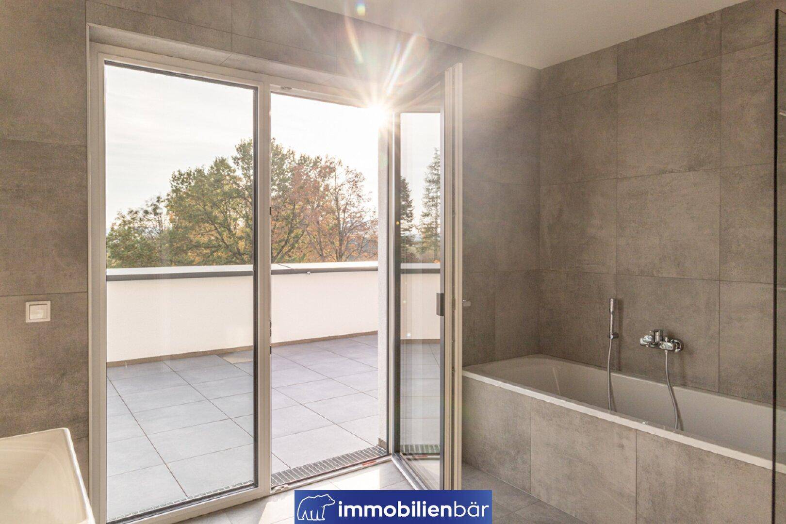 Modernes Badezimmer mit Badewanne, grauen Fliesen und großen Fenstertüren zur Terrasse.