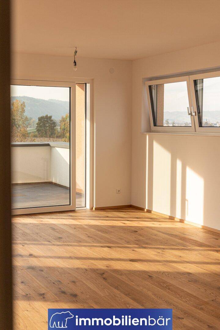 Sonniger Raum mit Holzboden, Fenster und Fenstertür, die auf eine Terrasse führt.