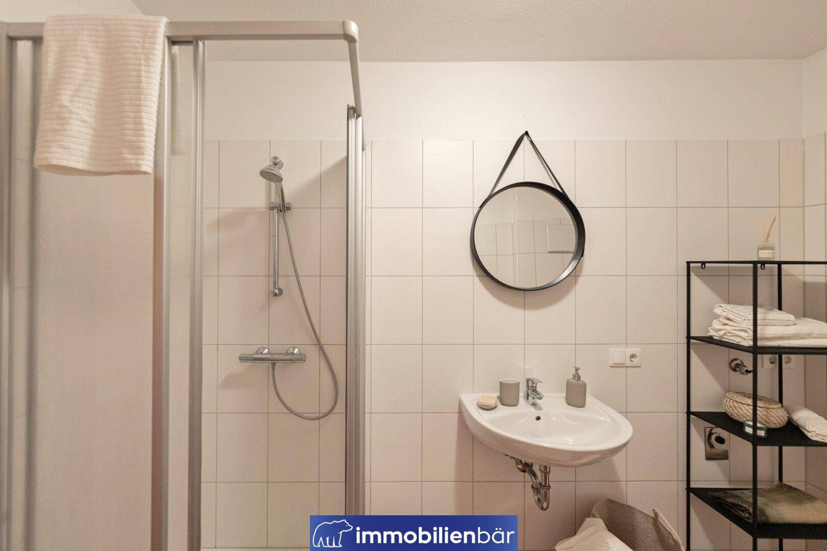 Badezimmer mit Duschkabine, Waschbecken und rundem Spiegel, hell gefliest und gut beleuchtet.