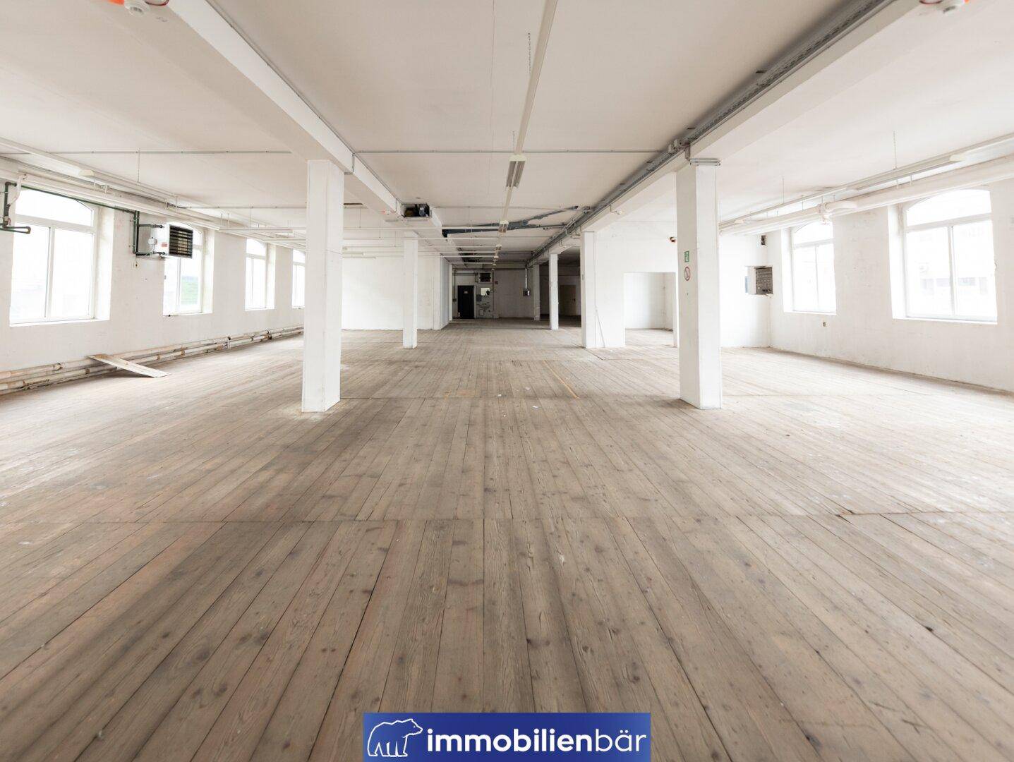 Großer, leerer Lagerraum mit Holzboden, weißen Wänden und sichtbaren Deckeninstallationen, ideal für industrielle Nutzung.