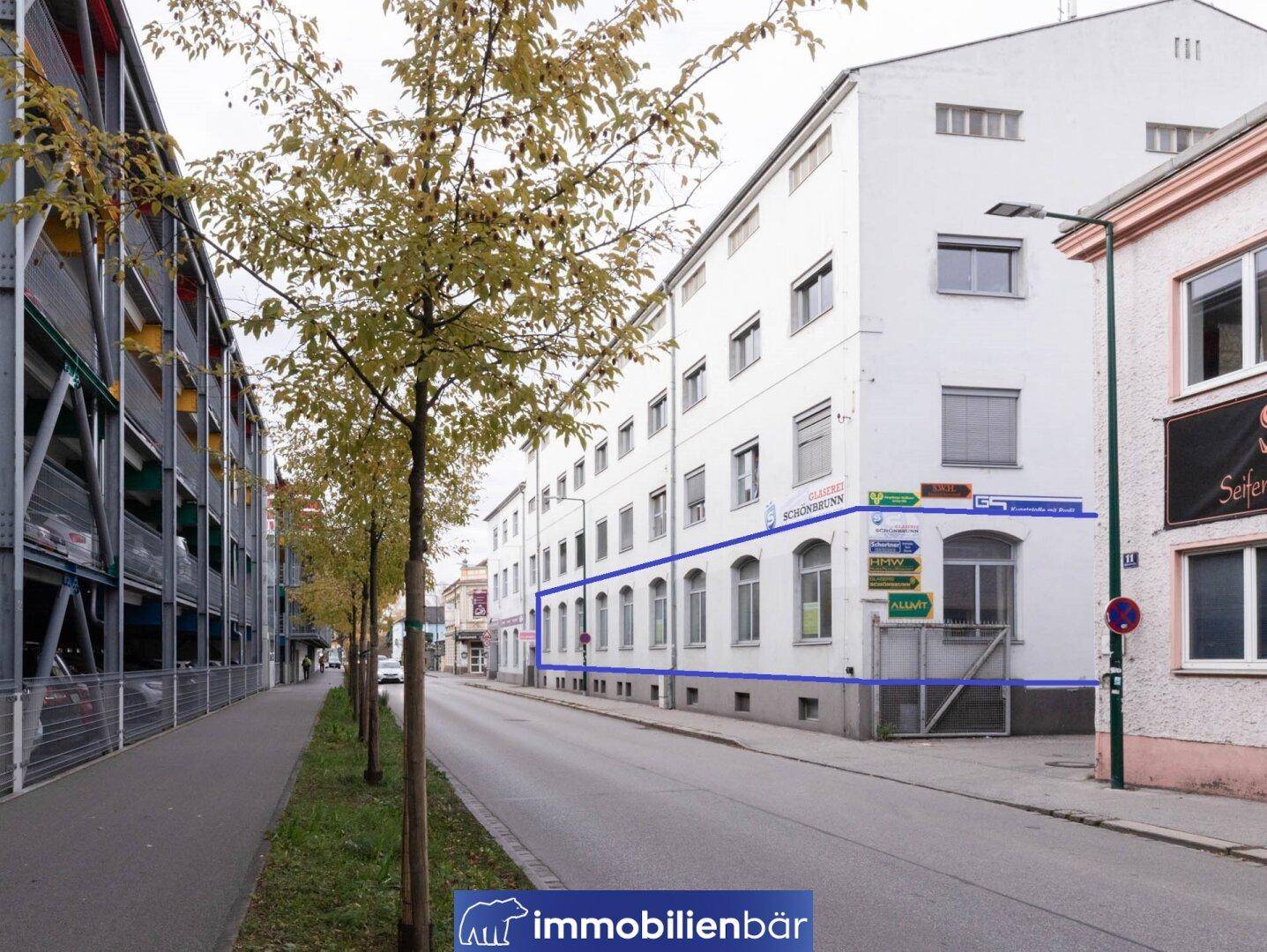 Straßenansicht des Gebäudes mit Erdgeschossfenstern und einem angrenzenden mehrstöckigen Parkhaus.