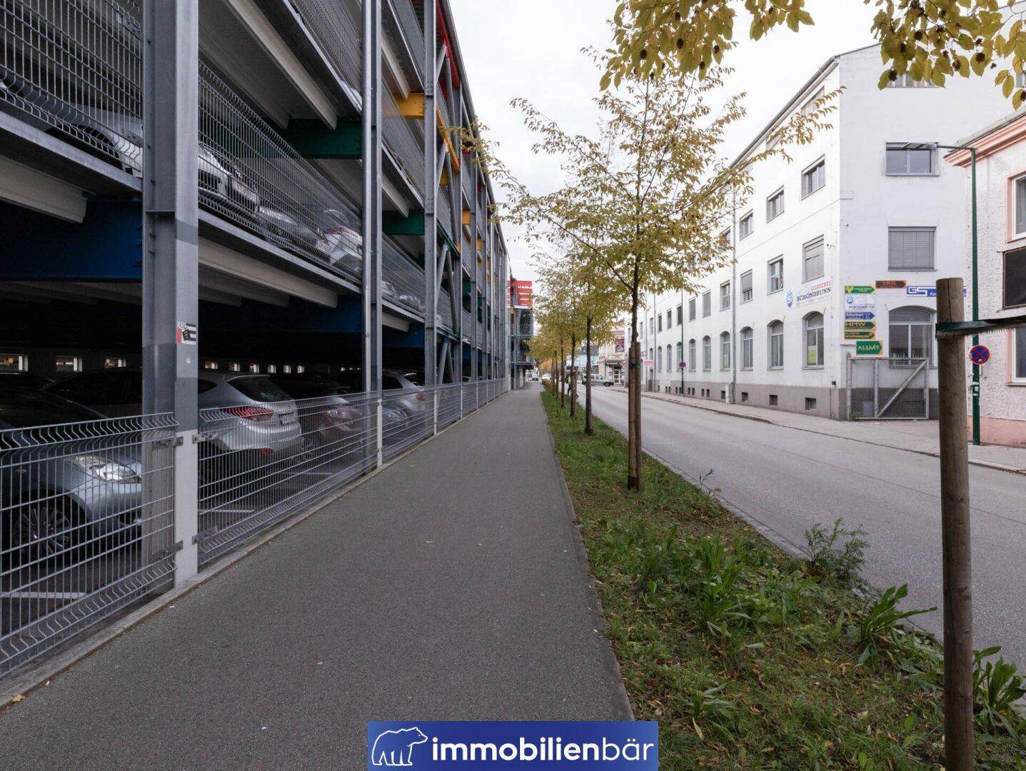 Straßenansicht mit einem mehrstöckigen Parkhaus auf der linken Seite und einer Baumreihe entlang der Straße.