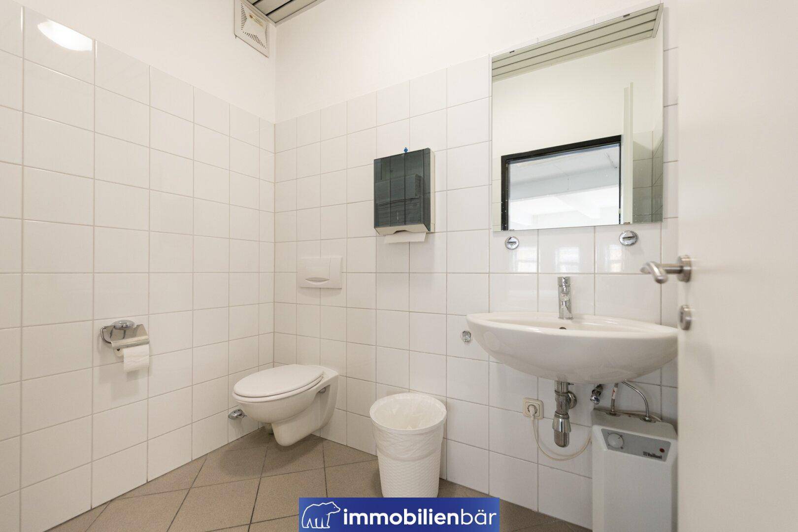 Einfache Toilette mit weißen Fliesen an den Wänden, einem Waschbecken, WC und einem Spiegel.