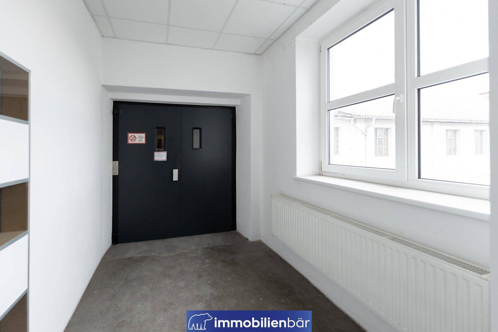 Geschlossene Aufzugstür neben einem Fenster und einem Heizkörper, der Boden ist aus Beton.
