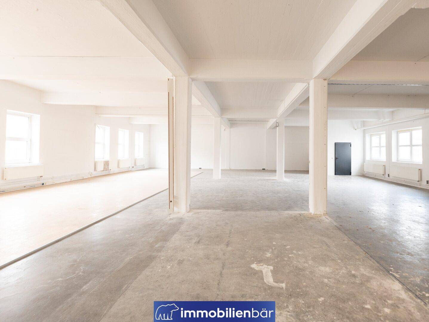 Heller, offener Büroraum mit Betonboden, weißen Wänden und tragenden Säulen, eine dunkle Tür ist im Hintergrund sichtbar.