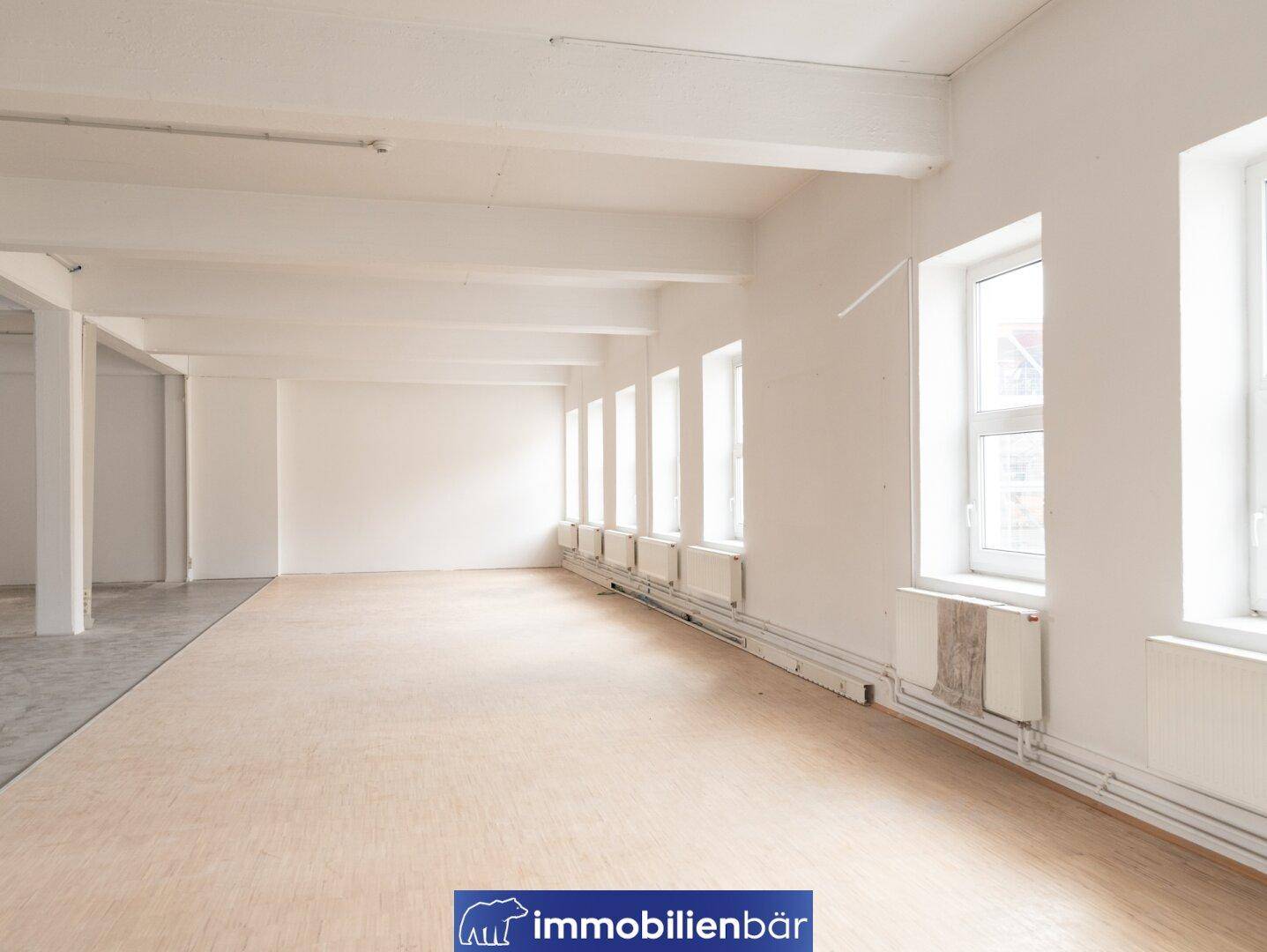 Geräumiger, heller Bürobereich mit hellem Laminatboden, weißen Wänden und einer Reihe von Fenstern mit Heizkörpern.