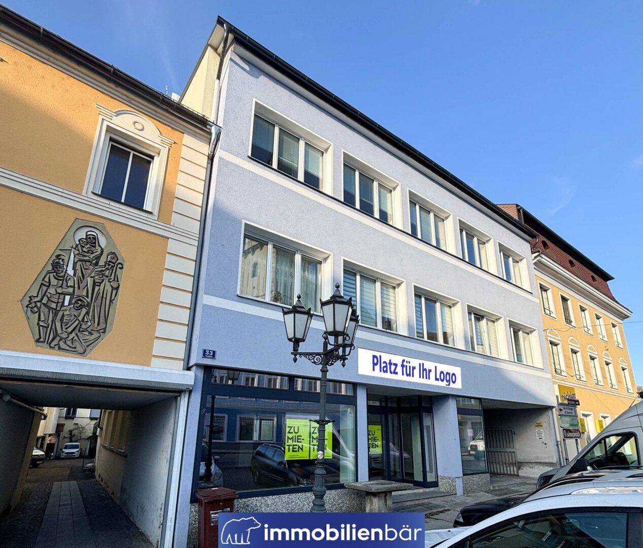 Mehrstöckiges Gebäude mit Geschäftsflächen im Erdgeschoss und Wohnungen darüber, umgeben von historischen Gebäuden in einer Stadtstraße.