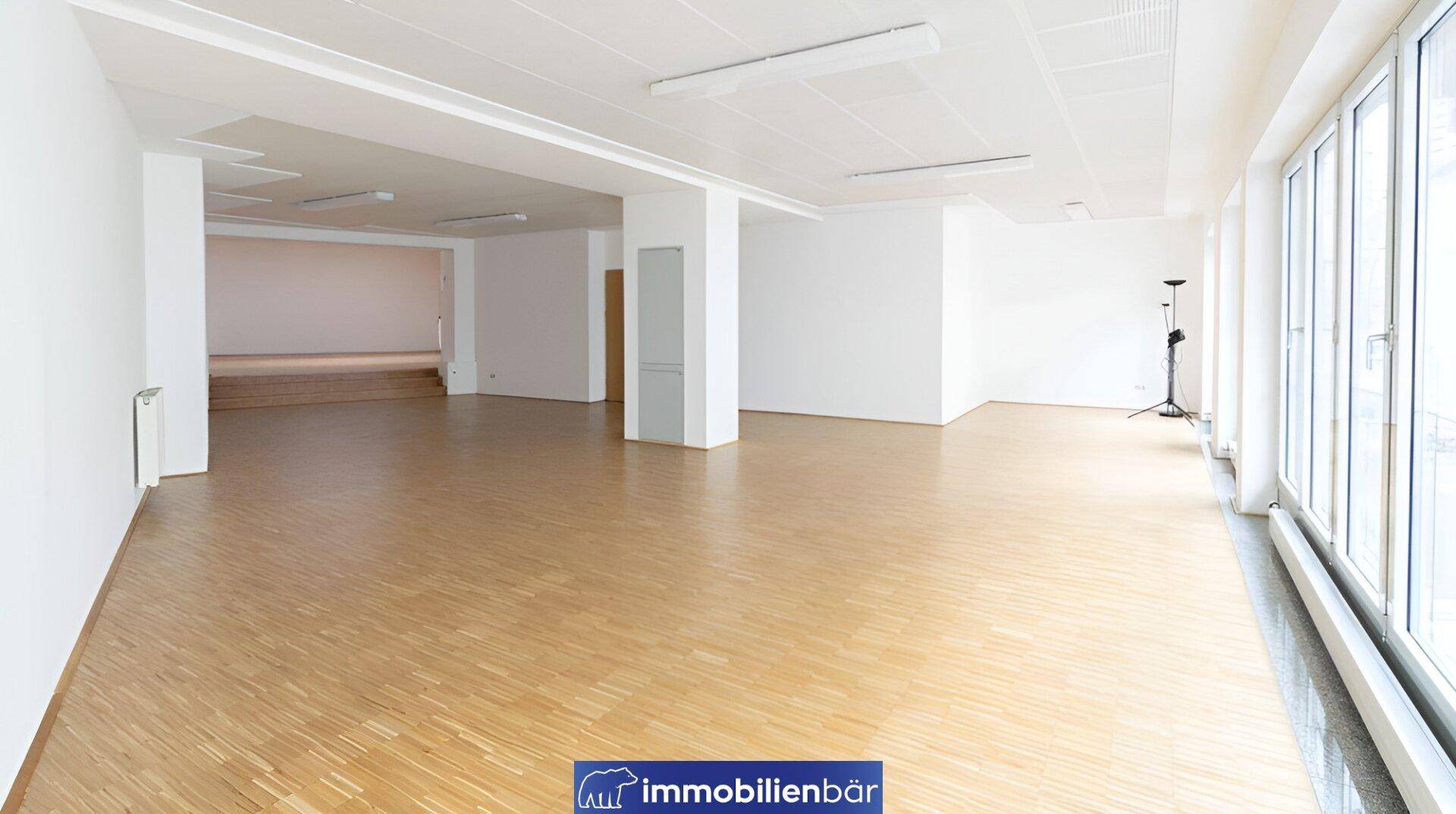 Heller Bürobereich mit Holzparkett und großen Fensterfronten, die viel Tageslicht hereinlassen.