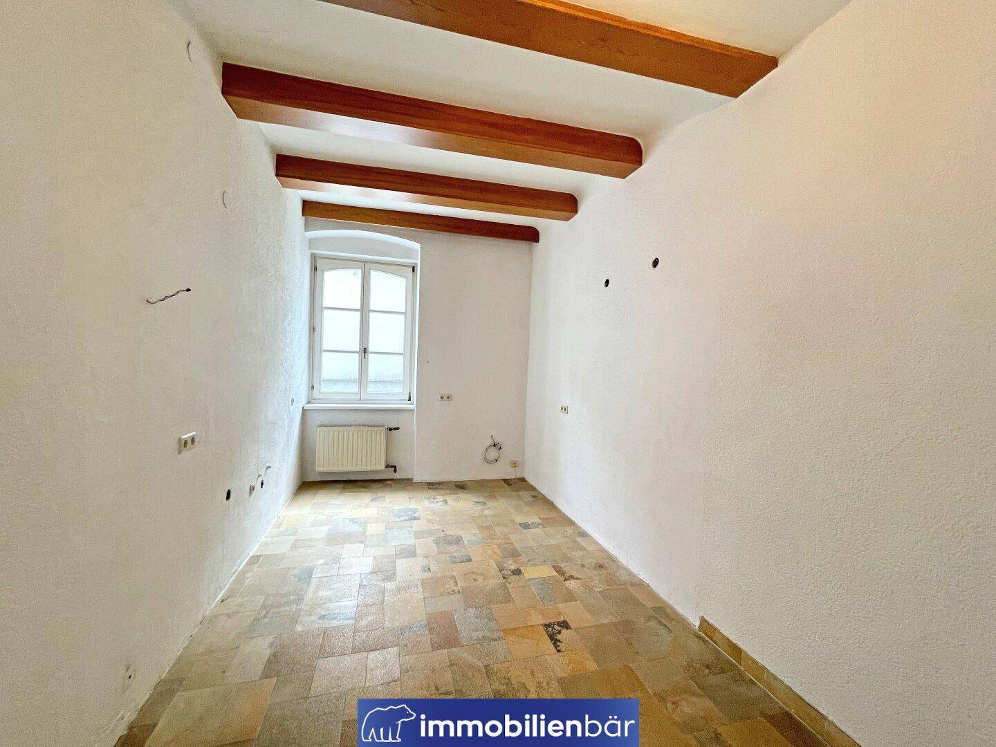 Raum mit sichtbaren Holzbalken, Fliesenboden und einem Fenster, renovierungsbedürftig.