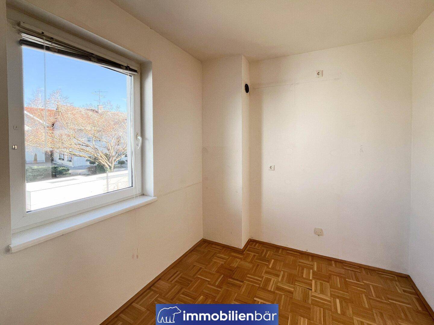 Helles Zimmer mit Parkettboden und einem großen Fenster, das viel Tageslicht hereinlässt.