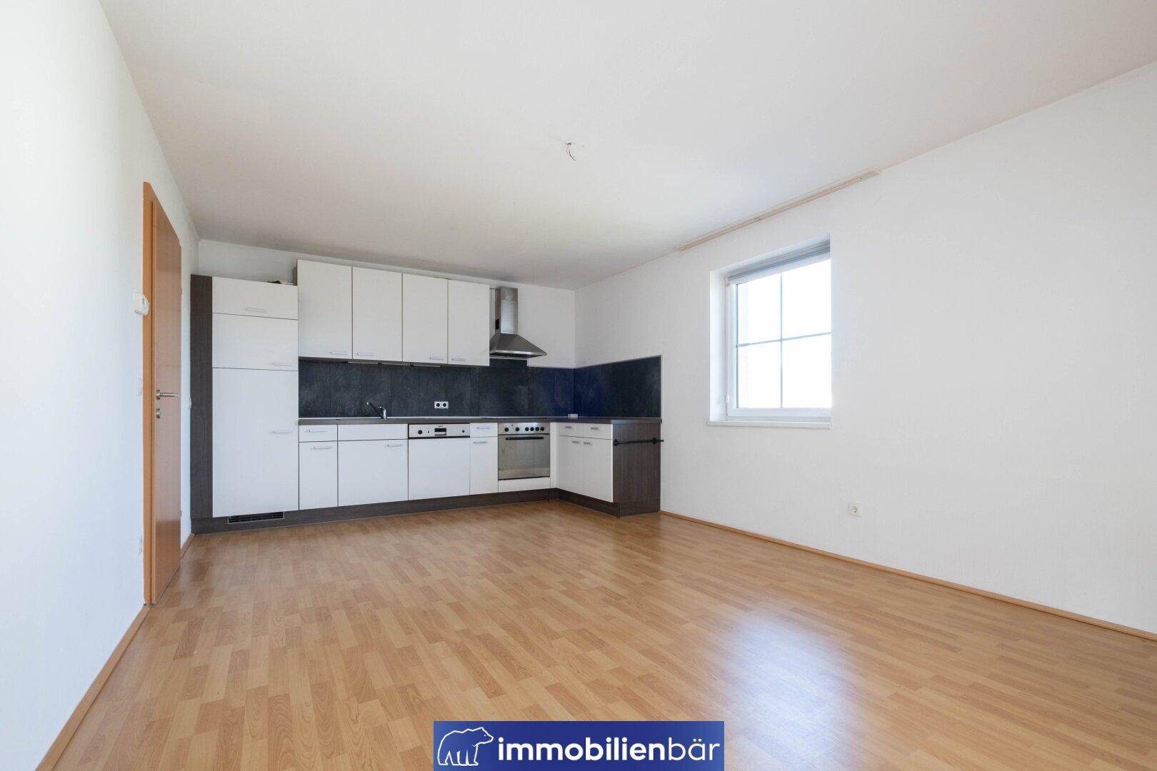 Offene Küche mit weißen Schränken, dunkler Arbeitsplatte und Laminatboden in einem hellen Raum.