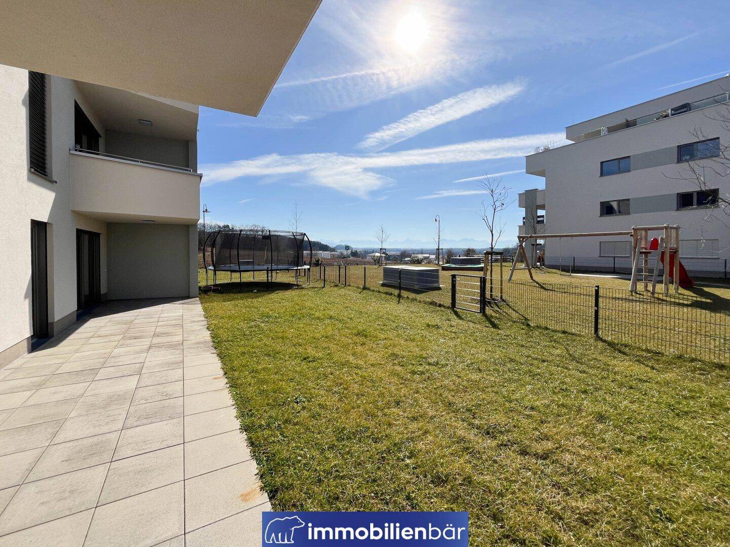 Sonniger Gemeinschaftsgarten mit Spielplatz und Trampolin neben dem modernen Wohngebäude.